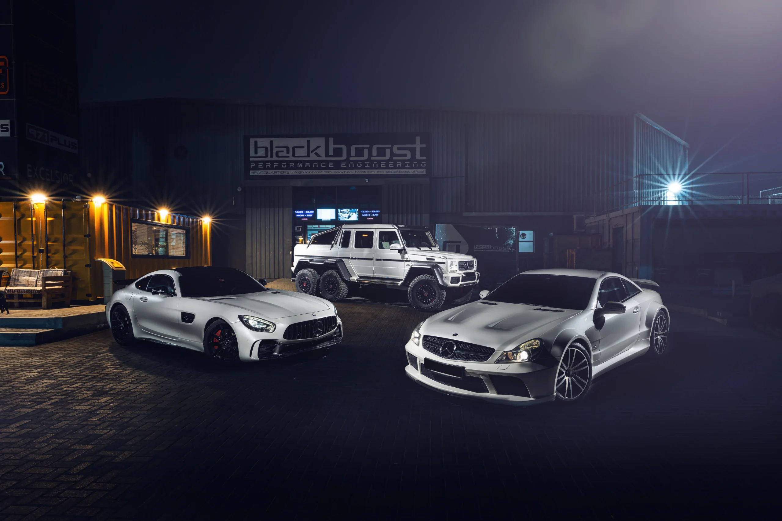 Three customized Mercedes-Benz performance vehicles, including two silver sports cars and a white G-Class SUV, parked outside Blackboost Performance Engineering workshop at night.